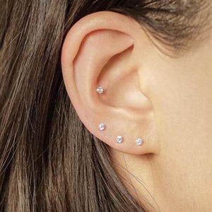 Könnte beinhalten: Nahaufnahme eines Ohrs mit vier kleinen, runden, durchsichtigen Ohrsteckern. Die Ohrringe sind gleichmäßig am Knorpel des Ohrs angeordnet. Der Hautton ist hell, und das Haar ist dunkelbraun.