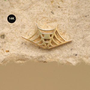 Op de afbeelding: Een goudkleurige hanger met een geometrisch ontwerp, dat lijkt op een gestileerd web of architecturaal element. De hanger is gemarkeerd met "14K" in een zwarte cirkel. Het geheel is geplaatst tegen een getextureerde, lichtbruine achtergrond.