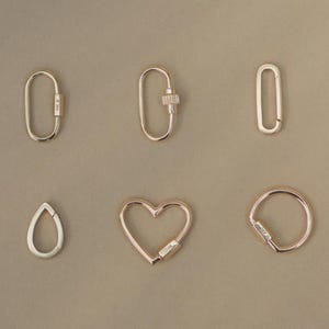 May include: Six gold-colored carabiner clasps in various shapes: oval, teardrop, heart, and round. Each clasp has a hinged opening and a secure closure. The clasps are displayed on a neutral, light brown background.