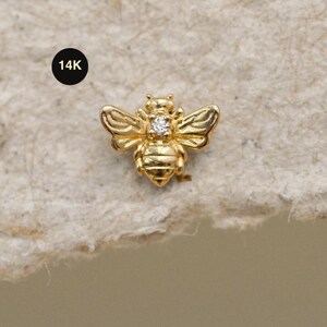 May include: A gold-colored bee-shaped stud earring with a small, clear gemstone in the center. The earring is marked with "14K" in a black circle. The bee has detailed wings and body segments.