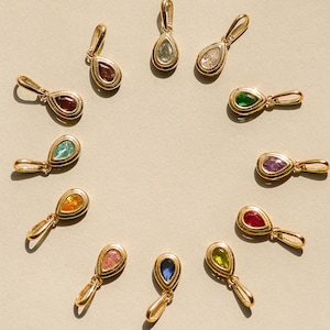 May include: A collection of eleven gold-toned pendants arranged in a circle, each featuring a teardrop-shaped gemstone in various colors including red, green, blue, yellow, pink, and clear, with a small gold loop at the top for attachment.