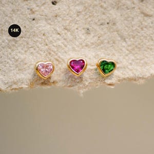 Puede incluir: Tres pendientes de oro con gemas en forma de corazón en rosa, magenta y verde. Los pendientes están engarzados en oro y se muestran sobre una superficie texturizada de color blanquecino. Un círculo negro con el texto "14K" está en la esquina superior izquierda.