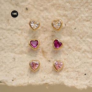 Può includere: Sei orecchini a lobo a forma di cuore con montatura in oro. Gli orecchini presentano pietre preziose trasparenti, rosa e magenta. Gli orecchini sono esposti su una superficie strutturata marrone chiaro. L'immagine include un piccolo cerchio nero con la scritta "14K".