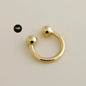 May include: A gold-colored, horseshoe-shaped earring with a ball at each end. The earring is marked with "14K" in a black circle. The jewelry is set against a neutral background.