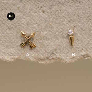 Könnte beinhalten: Zwei Schmuckstücke aus 14K Gold, beschriftet mit A und B. Stück A besteht aus zwei gekreuzten Pfeilen. Stück B ist ein einzelner Ohrstecker mit einem kleinen, klaren, rund geschliffenen Edelstein über einem glatten, kegelförmigen Goldanhänger.