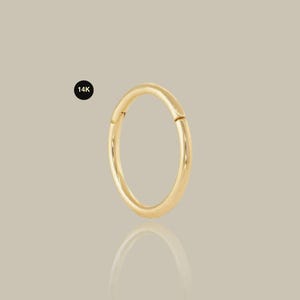 May include: A gold-colored, circular earring with a hinged clasp. The earring is smooth and shiny, reflecting light. A small black circle with the text "14K" is in the upper left corner. The earring is displayed on a neutral background.