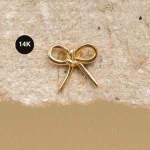 Può includere: Un piccolo gioiello dorato a forma di fiocco. Il fiocco è centrato su una superficie strutturata e beige. Un cerchio nero con la scritta "14K" si trova nell'angolo in alto a sinistra. Il design delicato del fiocco suggerisce che si tratti di un orecchino o di un ciondolo.