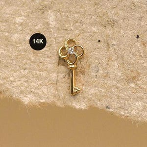 May include: A small, gold key pendant featuring a four-leaf clover design with a central diamond. The key is marked "14K" in a black circle. The pendant is set against a textured, tan background. A delicate piece of jewellery.
