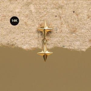 May include: A pair of gold-colored star-shaped earrings. The earrings are connected by a small chain, with one star above the other. The earrings are set against a textured, light brown background. The text "14K" is visible.