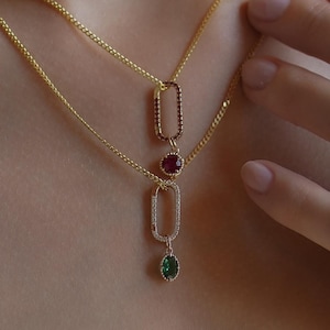 May include: A layered gold chain necklace featuring two elongated oval links. The top link is accented with small red stones, followed by a round red stone charm. The bottom link is encrusted with clear stones and holds a single oval green stone charm.