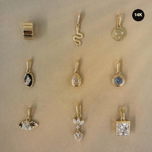 May include: A collection of nine 14K gold jewelry charms including a ribbed bead, a snake, a round disc with three diamonds, an oval black onyx piece, a pear-shaped stone, a blue round stone, a sunburst, a heart-shaped drop, and a square stone.