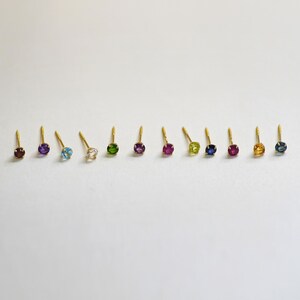 May include: A set of 10 gold stud earrings with different colored gemstones. The gemstones are arranged in a row from left to right: blue, purple, light blue, white, green, purple, blue, red, yellow, and blue.