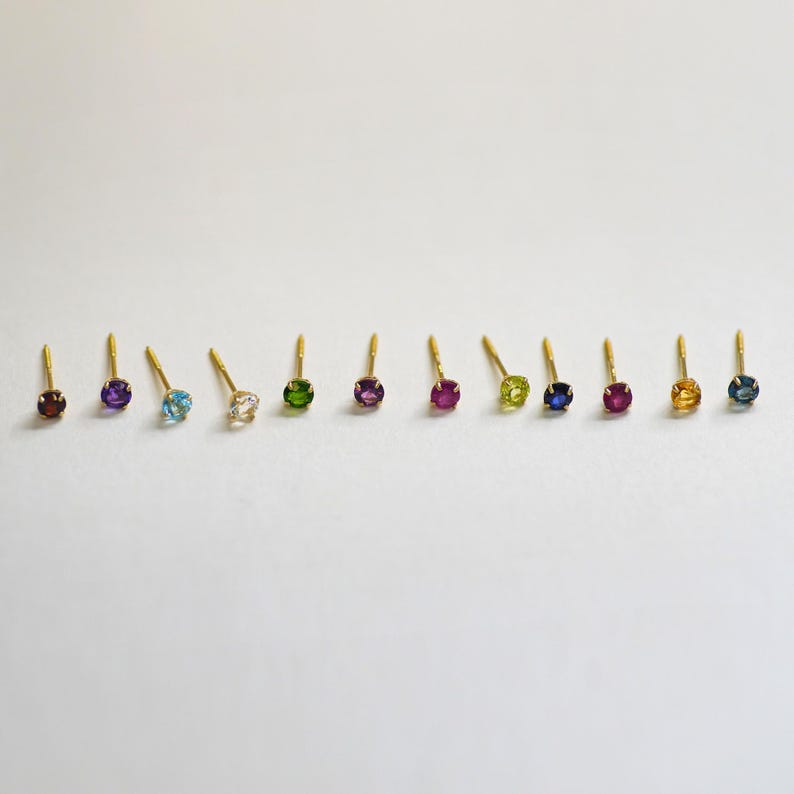 May include: A set of 10 gold stud earrings with different colored gemstones. The gemstones are arranged in a row from left to right: blue, purple, light blue, white, green, purple, blue, red, yellow, and blue.