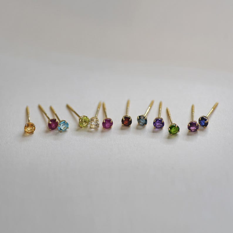 May include: A set of 10 gold stud earrings with different colored gemstones. The gemstones are: yellow, pink, blue, green, white, red, garnet, purple, and blue.