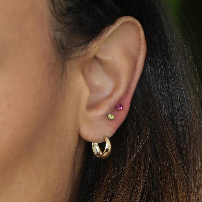 May include: A person's ear with a gold hoop earring and two small gemstone studs. The studs are pink and green.