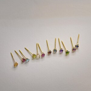 May include: A set of 12 gold stud earrings with different colored gemstones. The gemstones are set in a bezel setting and are all the same size. The earrings are arranged in a row on a white background.