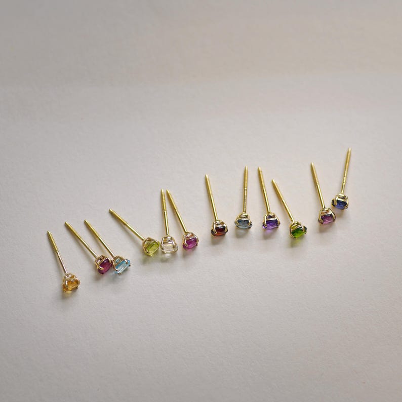 May include: A set of 12 gold stud earrings with different colored gemstones. The gemstones are set in a bezel setting and are all the same size. The earrings are arranged in a row on a white background.