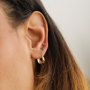 May include: A person wearing a gold hoop earring and two small gemstone earrings in their ear. The earrings are green and orange.
