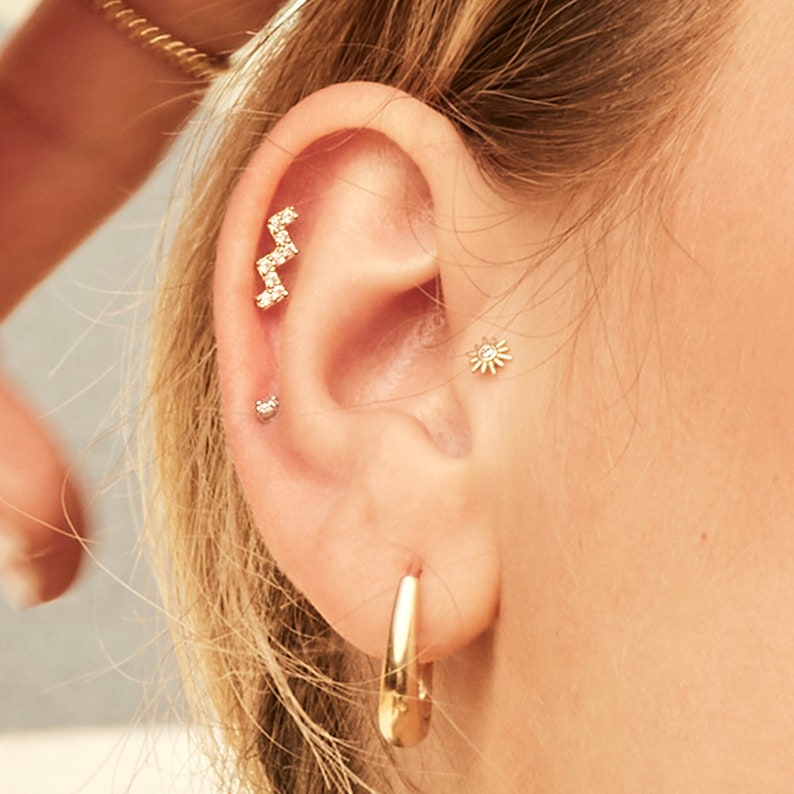 K&ouml;nnte beinhalten: Nahaufnahme eines Ohrs mit mehreren goldenen Ohrringen. Die Ohrringe umfassen einen kleinen Diamanten-Stecker, einen gr&ouml;&szlig;eren Diamanten-Stecker in Form einer Blume und einen langen, d&uuml;nnen Stab-Ohrring mit kleinen Diamanten, die in einem Zickzack-Muster gesetzt sind.