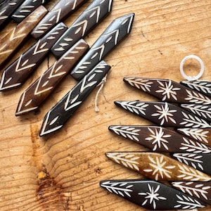 May include: A collection of handcrafted wooden decorative pieces in various shades of brown, adorned with white geometric and floral patterns. Some pieces are rectangular with arrow-like designs, while others are leaf-shaped with starburst motifs.