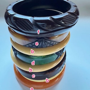 May include: A stack of six vintage bangles in various colors. The bangles include shades of orange, brown, green, and cream. The top bangle is dark brown with a carved leaf design. These bracelets are likely made of Bakelite or a similar material.