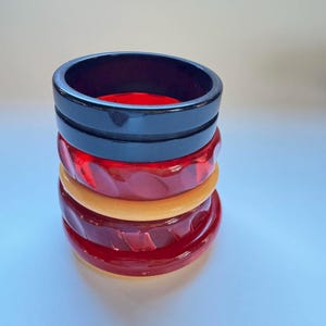 May include: A stack of four colorful bangles. The top bangle is black, followed by a red one, a tan one, and a final red bangle at the bottom. The red bangles have a textured, carved design. The bangles are stacked on a light blue surface.