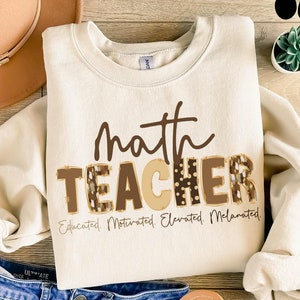 May include: A beige sweatshirt with the text "math teacher" in brown letters with a leopard print design. The text "Educated, Motivated, Elevated, Melamated" is printed below the main text.