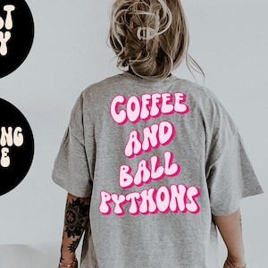 Peut inclure: Un t-shirt gris avec le texte "COFFEE AND BALL PYTHONS" imprimé en rose au dos. Le t-shirt est ample et a un col rond.