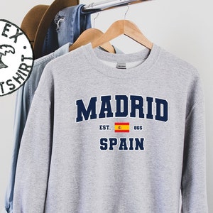 May include: A gray sweatshirt with the text "MADRID EST. 865 SPAIN" in navy blue lettering. The Spanish flag is printed between "EST." and "865".