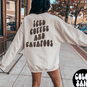 May include: A beige sweatshirt with the text "ICED COFFEE AND CAVAPOOS" in brown letters. The model is wearing the sweatshirt and white trainers. The text "COLOR SAND" and "WEARING LARGE" are written in white on black circles.