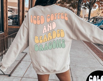 May include: A light beige sweatshirt with the text "ICED COFFEE AND BEARDED DRAGONS" in colourful, retro-style lettering. The model is wearing the sweatshirt and white trainers. The text "COLOR SAND" and "WEARING LARGE" are visible in the bottom right corner of the image.