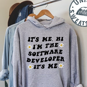 May include: Gray unisex sweatshirt with a black graphic that reads "IT'S ME, HI *I'M THE SOFTWARE DEVELOPER IT'S ME*" with white daisy flowers around the text. The sweatshirt has a hood and a kangaroo pocket.