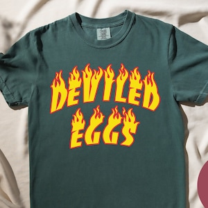 May include: A dark green t-shirt with the text "DEVILLED EGGS" in yellow and red letters with flames around the letters. The shirt is on a white fabric background.