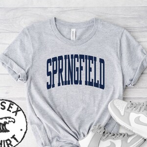 以下が含まれることがあります： グレーのユニセックスTシャツに、ネイビーブルーで「SPRINGFIELD」とプリントされています。Tシャツは、グレーとホワイトのハイカットスニーカーと合わせています。