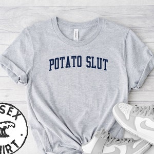 Pode incluir: Camiseta unisex cinza com o texto "POTATO SLUT" impresso em azul marinho. A camiseta é mostrada em uma superfície de madeira com dois tênis brancos.