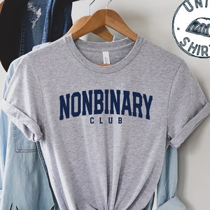 May include: A grey t-shirt with the navy blue text "NONBINARY CLUB". The shirt is displayed on a wooden hanger. A circular graphic with the words "UNISEX SHIRT" is visible in the top right corner of the image.