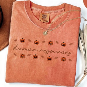 May include: A coral-colored t-shirt with the words "human resources" and jack-o'-lanterns. The shirt has a crew neck and short sleeves. A gold necklace and a brown hat are next to the shirt. The shirt is labeled "Comfort Colors".