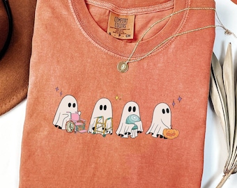 May include: A burnt orange t-shirt with a graphic of four ghosts carrying various items. The ghosts are white with black eyes and mouths. The text "Comfort Colors" is on a white tag attached to the shirt.