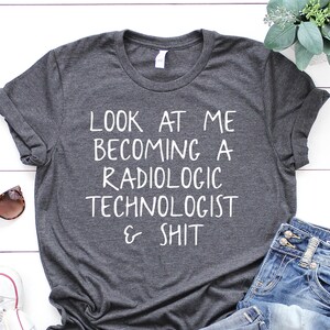 May include: A dark gray t-shirt with the text "LOOK AT ME BECOMING A RADIOLOGIC TECHNOLOGIST & SHIT" printed in white.