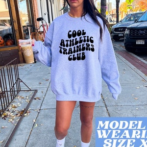 May include: A gray sweatshirt with the text "Cool Athletic Trainers Club" in black, retro-style lettering. The model is wearing a size XL.
