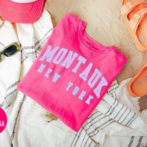 May include: A pink t-shirt with the text "Montauk New York" in blue lettering. The shirt is folded and lying on a white and gray striped towel with a pink hat and sunglasses. There are also two orange sandals on the sand.