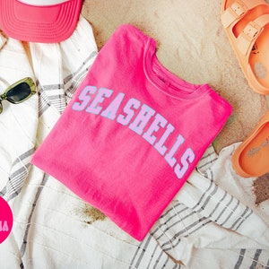 May include: A pink t-shirt with the text "SEASHELLS" in a light blue color. The t-shirt is folded and lying on a white and gray striped towel. There is a pink baseball cap, green sunglasses, and two orange sandals in the image.