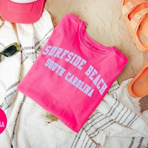 May include: Pink t-shirt with the text "Surfside Beach South Carolina" in purple lettering. The t-shirt is folded and lying on a white and gray striped towel. There are orange sandals, sunglasses, and a pink hat in the background.