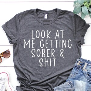 May include: A dark gray t-shirt with the text "LOOK AT ME GETTING SOBER & SHIT" printed in white on the front. The shirt is laid out on a white wooden surface with a pair of white sneakers, a pair of sunglasses, and a pair of blue jeans.