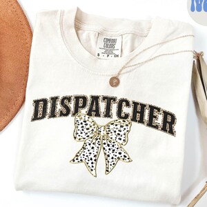 May include: A cream colored t-shirt with the word "DISPATCHER" in black outlined in gold glitter. A black and white leopard print bow is below the word. The shirt is on a white background.