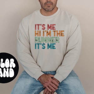 May include: A light beige sweatshirt with the text "It's me Hi I'm the runner It's me" in rainbow colors. The text is in a retro font style.