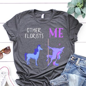 May include: A dark gray t-shirt with a humorous graphic of two unicorns. The text on the shirt reads "Other Florists" and "Me". The unicorn on the left is standing normally, while the unicorn on the right is doing a pole dance.