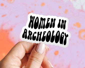 May include: A white sticker with black text that reads "Women in Archeology" in a retro font. The sticker is being held by a hand against a pink and orange background.