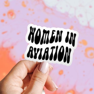 May include: A white sticker with black text that reads "Women in Aviation" in a retro font. The sticker is being held by a hand against a pink and orange background.