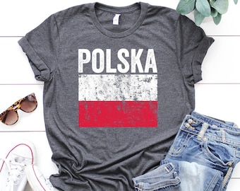 Peut inclure: Un t-shirt gris avec un drapeau polonais blanc délavé et le mot "POLSKA" imprimé dessus. Le t-shirt est posé sur une surface en bois blanc avec une paire de lunettes de soleil, une paire de baskets blanches et un jean bleu.